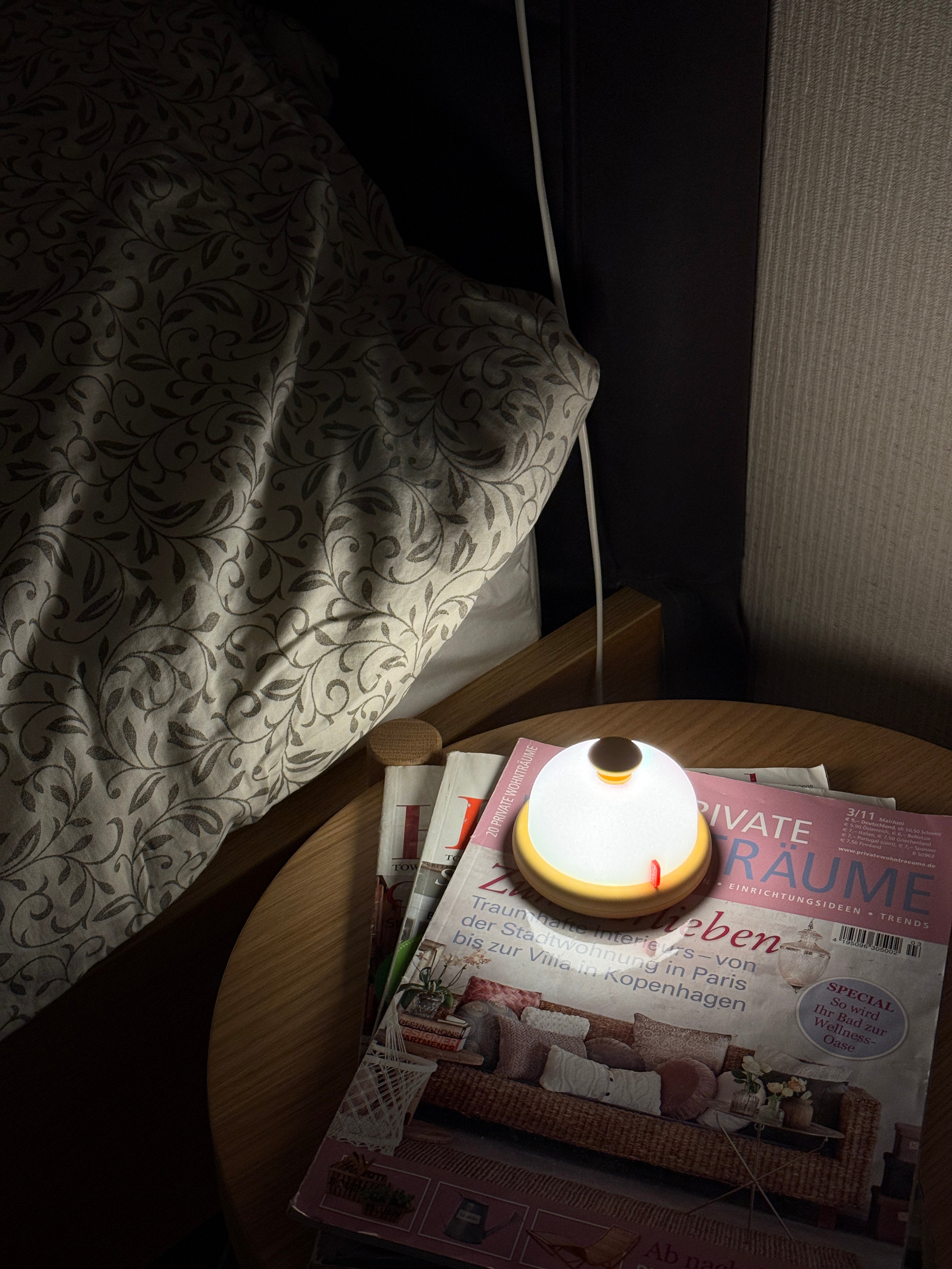 Small lamp on a table next to a bed with a magazine underneath