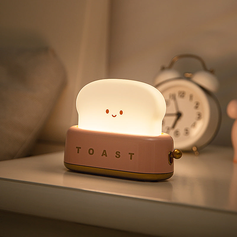 Pink night light shaped like a toaster with a smiling face on a surface, next to an alarm clock.